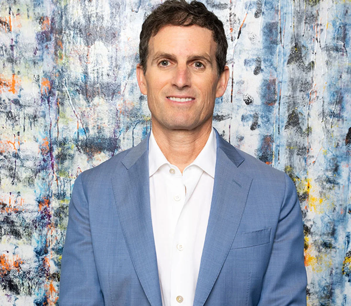 Portfolio shot of Dr. Rosenburg smiling man with short brown hair wearing a light blue blazer and white shirt, posing with arms crossed against a colorful backdrop. - Rhinoplasty in New York, NY